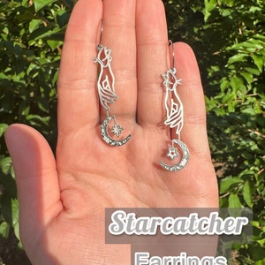 May include: Silver-toned Starcatcher earrings. Each earring features a hand silhouette holding a crescent moon with a star pendant. The earrings are displayed against a green leafy background. The text "Starcatcher Earrings" is in the image.