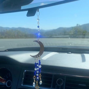 May include: A gold crescent moon charm with blue and clear crystal beads hanging from a rearview mirror in a car.