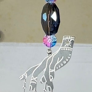 May include: A silver hand charm with a celestial theme. The hand is holding a crescent moon, a star, and a sun. The charm is adorned with iridescent blue, pink, and black beads.