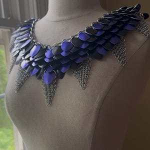 May include: A handcrafted, scale mail collar in shades of black and purple, displayed on a mannequin. The collar features overlapping metal scales and chainmail fringe, creating a unique, fantasy-inspired accessory.