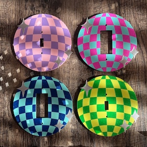 以下が含まれることがあります： ピンク、紫、ティール、青、黄色の市松模様のカラフルなライトスイッチカバー4枚。各カバーにはスイッチ用の長方形の開口部があり、シルバーの星のアクセントで装飾されています。