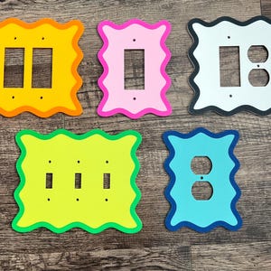 以下が含まれることがあります： カラフルな装飾的なライトスイッチカバーのコレクション。カバーは、黄色、ピンク、白、緑、青など、さまざまな色の波状の不規則なボーダーが特徴です。各カバーには、さまざまなスイッチ構成用の切り欠きがあります。