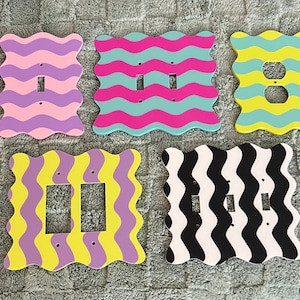 May include: Five decorative light switch covers with a wavy design. The covers feature a range of colours: pink and purple, pink and teal, yellow and teal, yellow and purple, and black and white. A fun and colourful home decor item.