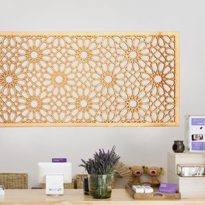 May include: A wooden wall decoration featuring a geometric pattern of interlacing circles and stars. The decor is light brown and has a natural wood finish. It is hanging on a white wall above a counter with a till, a basket, and a vase of lavender.