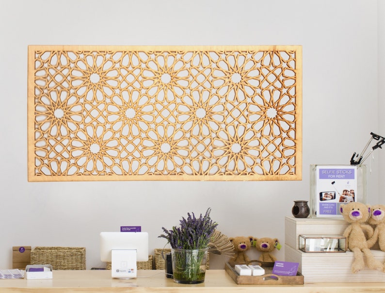 May include: A wooden wall decoration featuring a geometric pattern of interlacing circles and stars. The decor is light brown and has a natural wood finish. It is hanging on a white wall above a counter with a till, a basket, and a vase of lavender.