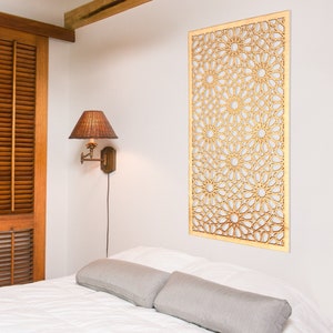 May include: A white bedroom with a wooden headboard and a decorative wooden wall panel with a geometric pattern. The wall panel is light brown and has an intricate design. There is a lamp on the wall and a white bed with two grey pillows.