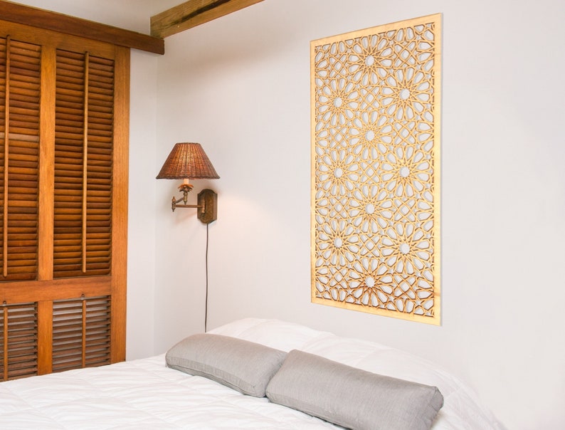 May include: A white bedroom with a wooden headboard and a decorative wooden wall panel with a geometric pattern. The wall panel is light brown and has an intricate design. There is a lamp on the wall and a white bed with two grey pillows.