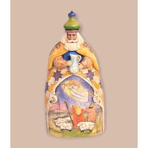 May include: A carved wooden figurine of a robed figure holding a pitcher. The figure has a white beard and a green and blue hat. The robe is yellow and patterned with purple and blue. The base depicts an angel, shepherds, and sheep.