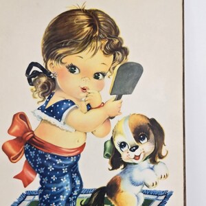 Puede incluir: Ilustración vintage de una niña en pijama azul con lunares blancos, sosteniendo un espejo, con un cachorro. La niña tiene cabello castaño rizado y un lazo rojo. El cachorro tiene pelaje marrón y blanco y un lazo verde.