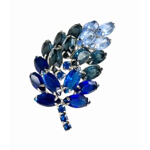 Könnte beinhalten: Eine Vintage-Brosche in Blattform mit einem Farbverlauf aus blauen Edelsteinen. Die Brosche hat eine silberfarbene Metalleinfassung und einen Stiel mit kleinen blauen Steinen. Die Edelsteine variieren von tiefem Marineblau bis Hellblau.