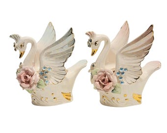 May include: Two ceramic swan ornaments with outstretched wings, cream-coloured base, gold accents, and delicate pink roses. The swans have detailed feathers and are adorned with blue and clear crystal accents.