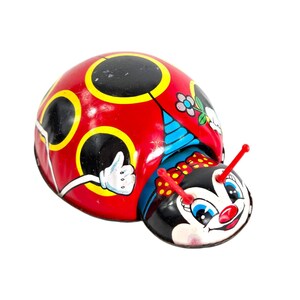 May include: A vintage, wind-up toy ladybug. The ladybug has a red body with black and yellow spots, a black head with a smiling face, and red antennae. A white gloved hand gives a thumbs up. The toy is made of metal.