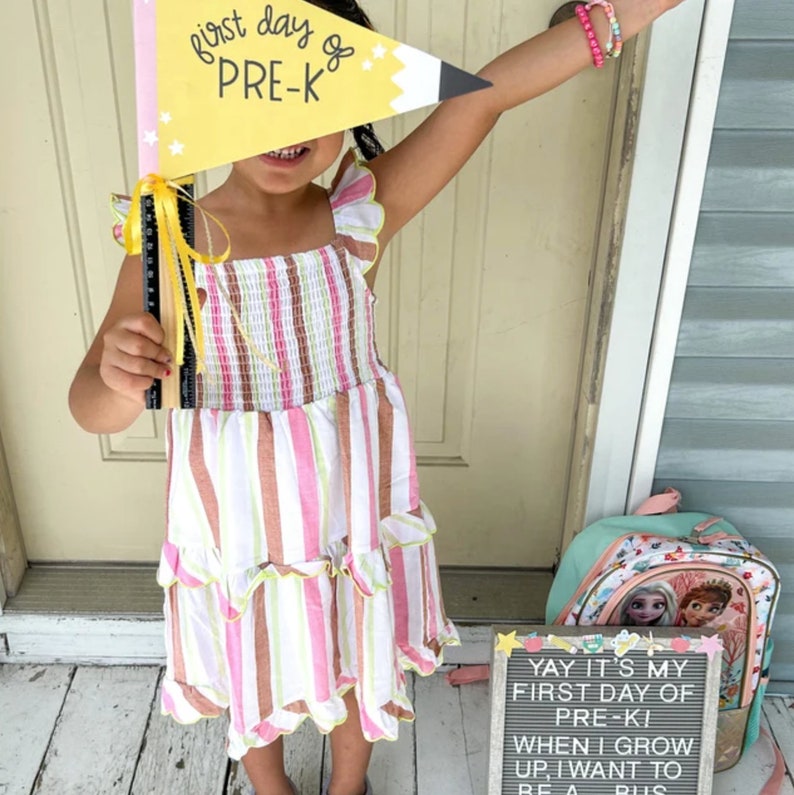 May include: A young girl wearing a pink and white striped dress holds a yellow and black pencil-shaped sign that says "First day of Pre-K". She is standing in front of a door with a green and pink backpack with a Frozen design next to her. The backpack has a letter board that says "Yay it's my first day of Pre-Ki when I grow up I want to be a bus"