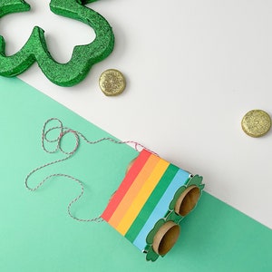 May include: A rainbow-colored cardboard box with a shamrock design and two empty toilet paper roll holders. The box is tied with a red and white string and is sitting on a green surface. There are gold coins scattered around the box.