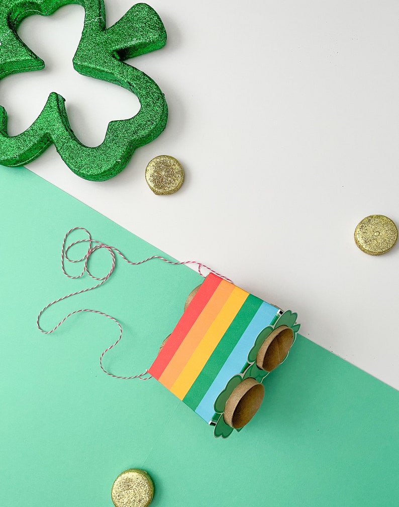 May include: A rainbow-colored cardboard box with a shamrock design and two empty toilet paper roll holders. The box is tied with a red and white string and is sitting on a green surface. There are gold coins scattered around the box.