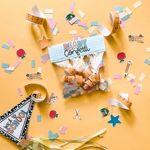 Pode incluir: Um saco de pl&aacute;stico transparente cheio de confete colorido, incluindo formas de &ocirc;nibus escolar, estrela e r&eacute;gua. O saco est&aacute; etiquetado como "Ready Confetti". O fundo &eacute; uma superf&iacute;cie amarela com confete espalhado.