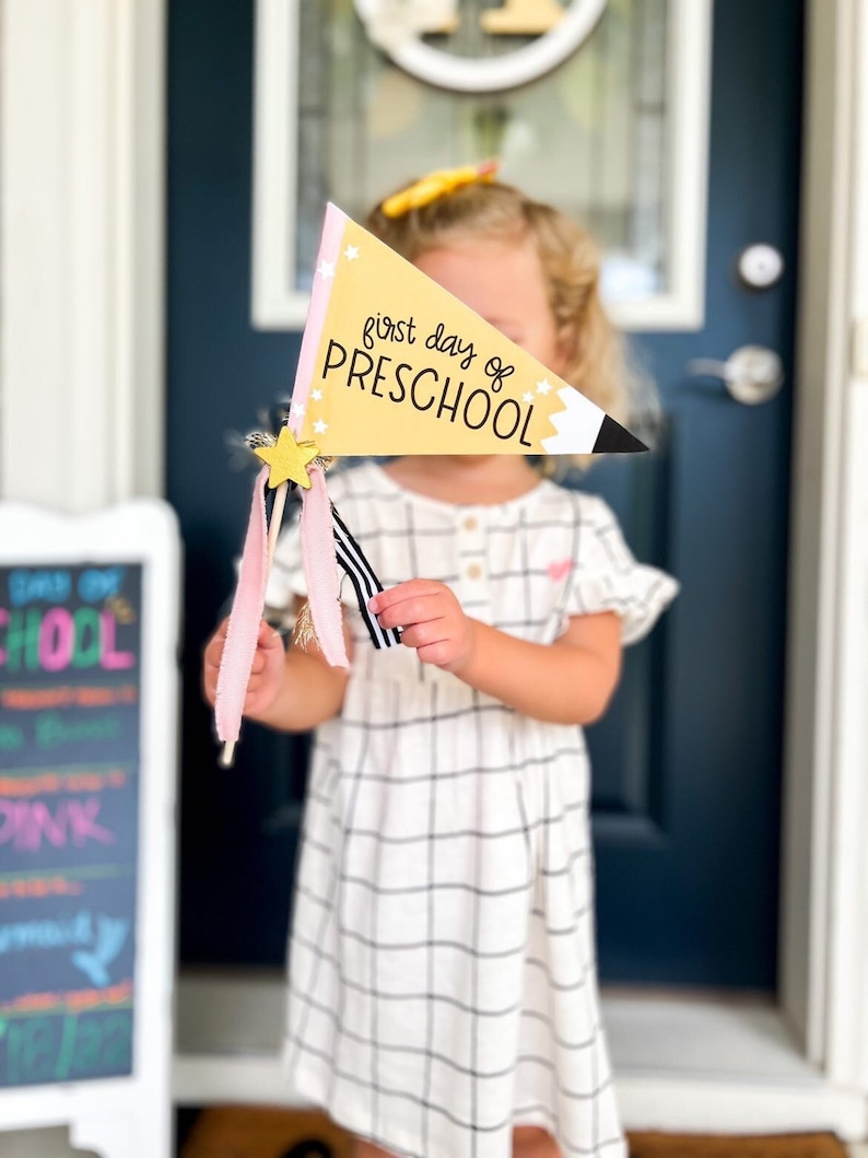 May include: A child holds a yellow and pink pennant with the text "first day of PRESCHOOL" and gold stars. The pennant is shaped like a pencil.