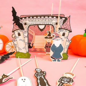 Può includere: Un set di topper per torte a tema Halloween con una strega, un mago, un fantasma, uno scheletro e un gatto nero. I topper sono realizzati in carta e sono progettati per assomigliare a personaggi di una fiaba. I topper sono posizionati su uno sfondo rosa.