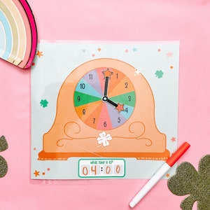 Puede incluir: Una esfera de reloj colorida con una manecilla de hora y una manecilla de minutos móviles. La esfera del reloj está dividida en 12 secciones, cada una con un color diferente. El reloj está configurado a las 4:00. El texto "¿qué hora es?" está impreso sobre la esfera del reloj. El reloj está colocado sobre un fondo rosa.