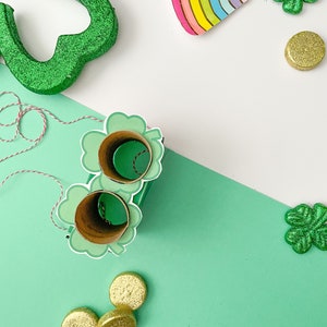 May include: A green shamrock cutout with glitter, a rainbow paper plate, and gold glitter coins on a white and green background. There are also two green shamrock cutouts with holes in the center, and a string of red and white twine.