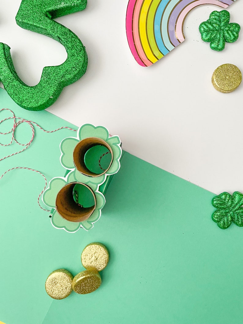 May include: A green shamrock cutout with glitter, a rainbow paper plate, and gold glitter coins on a white and green background. There are also two green shamrock cutouts with holes in the center, and a string of red and white twine.