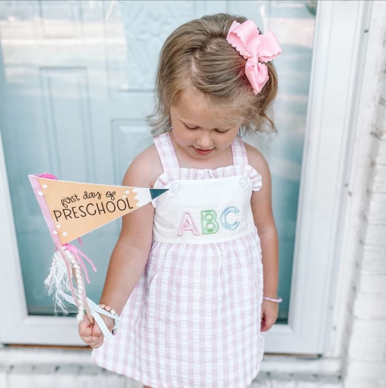 May include: A young girl in a pink and white gingham dress holds a sign that says "first day of preschool" with a pencil design. The sign is yellow with pink and blue accents. The girl has a pink bow in her hair.