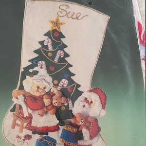 Könnte beinhalten: Ein weißer Filzstrumpf mit einem grünen Weihnachtsbaum, Figuren von Weihnachtsmann und Weihnachtsfrau und dem Namen "Sue" oben bestickt.