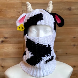 May include: A white and black crocheted cow-print balaclava with a yellow tag that says "916" and pink ears.