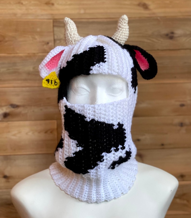 May include: A white and black crocheted cow-print balaclava with a yellow tag that says "916" and pink ears.