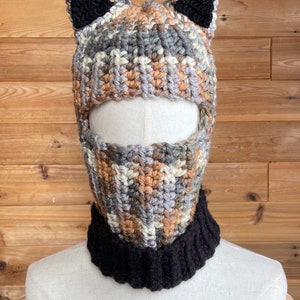 May include: A crocheted hat with cat ears. The hat is brown, gray, and beige with black ears. The hat has a face mask attached to it. The face mask is also crocheted and is brown, gray, and beige.