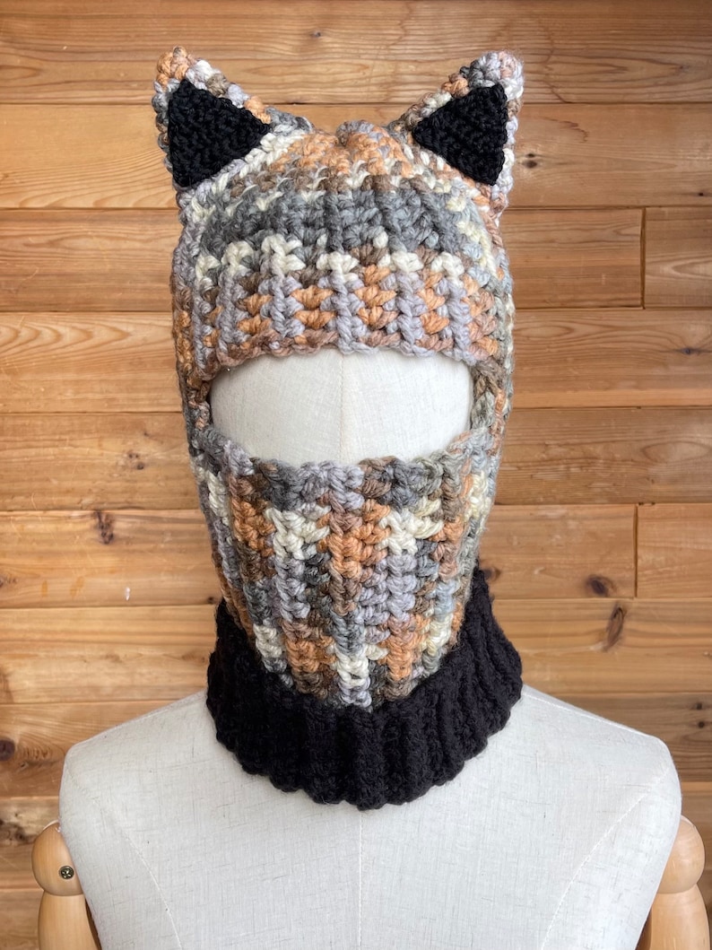 May include: A crocheted hat with cat ears. The hat is brown, gray, and beige with black ears. The hat has a face mask attached to it. The face mask is also crocheted and is brown, gray, and beige.
