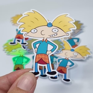 May include: A die-cut sticker depicting a cartoon character with blonde hair, a blue top, and a plaid skirt. The character is smiling and wearing a blue hat. Other stickers are visible in the background.