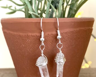 May include: A pair of clear quartz crystal earrings. Each earring features a clear crystal wrapped in silver-colored wire, suspended from a silver-colored hook. The earrings are displayed in front of a terracotta pot with a green plant.