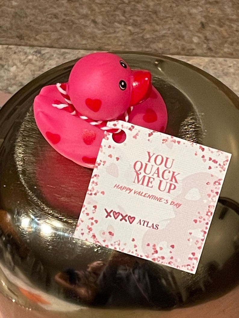 May include: A pink rubber duck with red heart designs and a white ribbon around its neck. The duck is sitting on a gold surface with a white card that says "You Quack Me Up Happy Valentine's Day xoxo Atlas".
