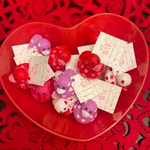 May include: A red heart-shaped bowl filled with rubber duckies decorated with pink and white hearts. Each duck has a tag attached that says "You Quack Me Up" with a heart and the text "Happy Valentine's Day".