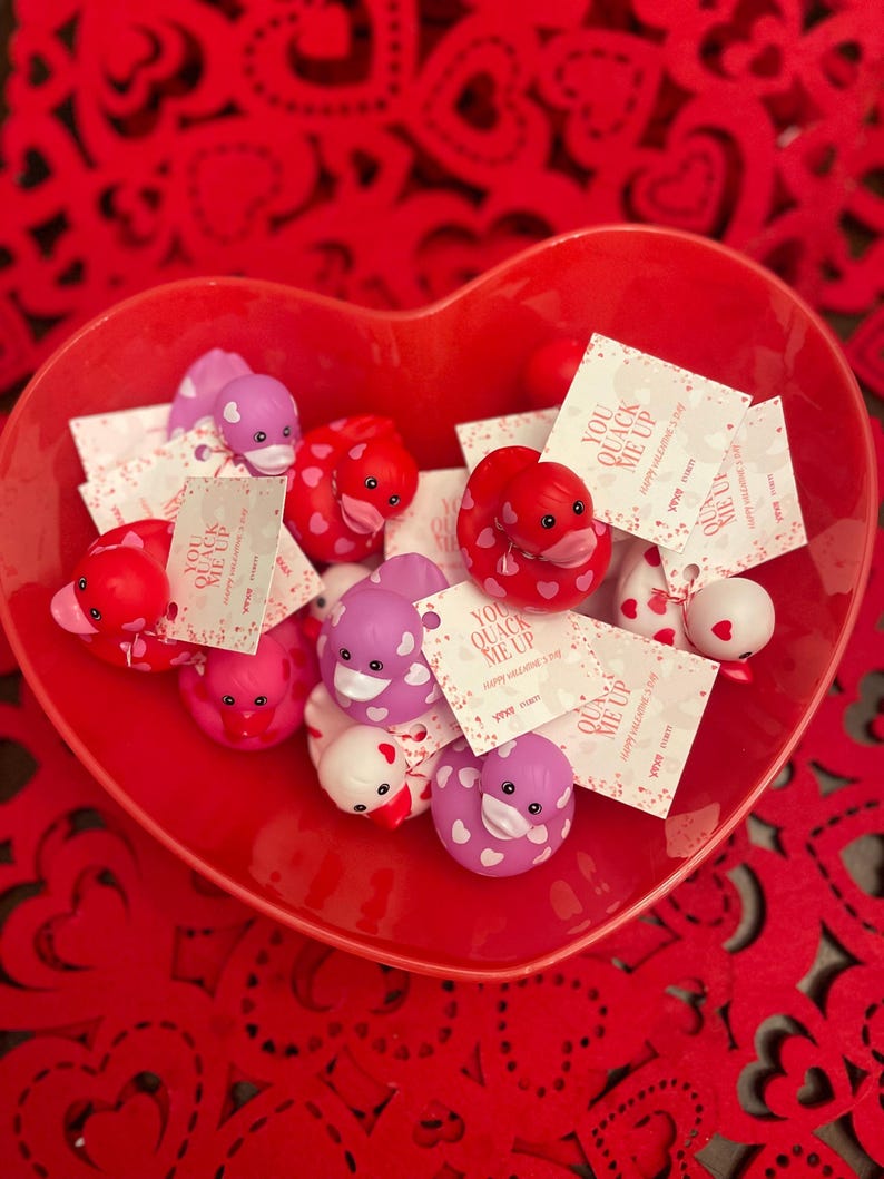 May include: A red heart-shaped bowl filled with rubber duckies decorated with pink and white hearts. Each duck has a tag attached that says "You Quack Me Up" with a heart and the text "Happy Valentine's Day".