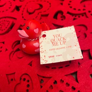 May include: A red rubber duck with pink accents and white polka dots sits on a red felt background with cut-out hearts. A white card with the text "You Quack Me Up Happy Valentine's Day XOXO Everett" is next to the duck.