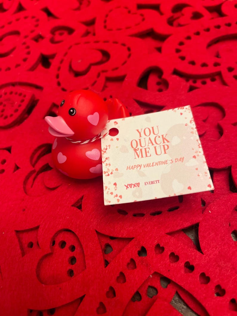 May include: A red rubber duck with pink accents and white polka dots sits on a red felt background with cut-out hearts. A white card with the text "You Quack Me Up Happy Valentine's Day XOXO Everett" is next to the duck.