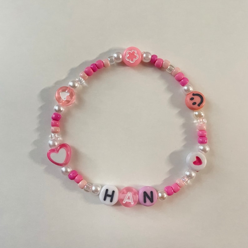 May include: Pink and white beaded bracelet with the name "HAN" spelled out in white letters with black outlines. The bracelet also features pink and white heart charms, a flower charm, and a smiley face charm.