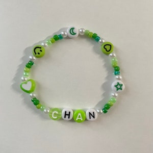 May include: Green and white beaded bracelet with the name "CHAN" spelled out in white letters on green beads. The bracelet also features a smiley face, a heart, a crescent moon, and a star.