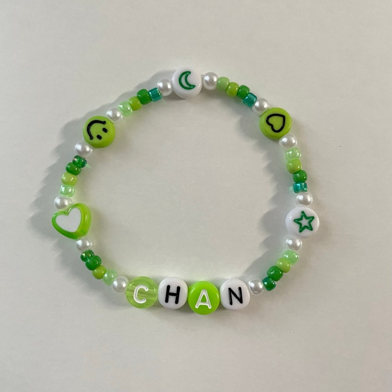 May include: Green and white beaded bracelet with the name "CHAN" spelled out in white letters on green beads. The bracelet also features a smiley face, a heart, a crescent moon, and a star.