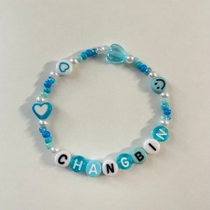 May include: A blue and white beaded bracelet with the name "CHANGBIN" spelled out in black letters. The bracelet also features a heart, a smiley face, and a clear heart charm.