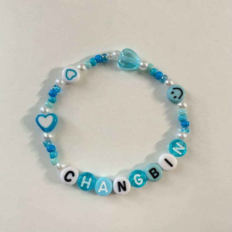 May include: A blue and white beaded bracelet with the name "CHANGBIN" spelled out in black letters. The bracelet also features a heart, a smiley face, and a clear heart charm.