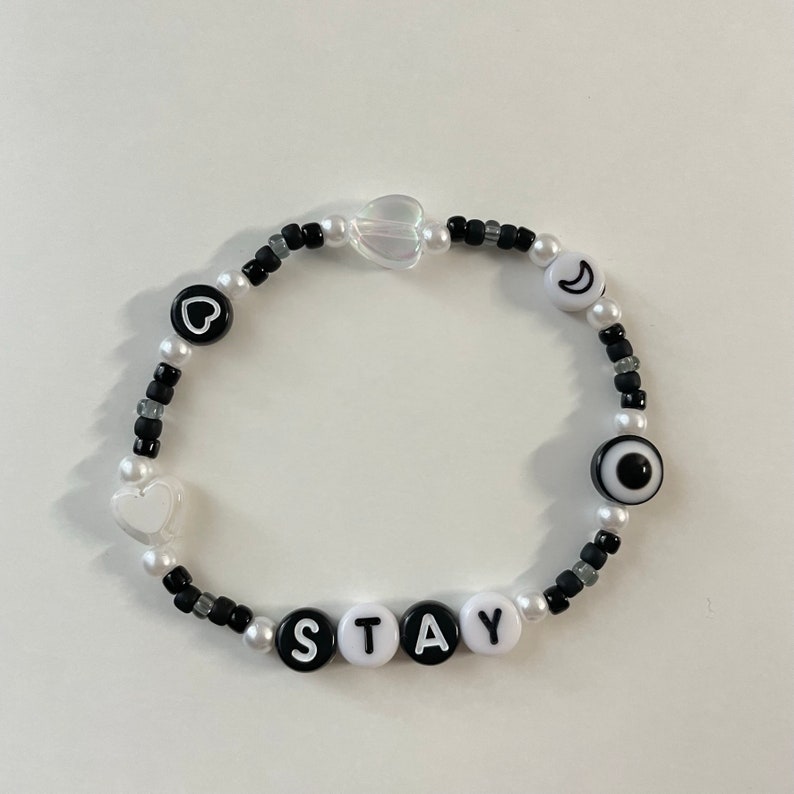 May include: A black and white beaded bracelet with the word "STAY" spelled out in white beads. The bracelet also features a heart, a crescent moon, and an evil eye charm.