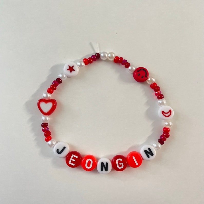 May include: A red and white beaded bracelet with the name "JEONGIN" spelled out in black letters. The bracelet also features a red heart, a red star, and a red smiley face.