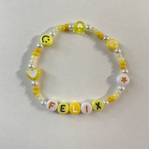 May include: A yellow and white beaded bracelet with the name "FELIX" spelled out in white letters on yellow beads. The bracelet also features a smiley face, a heart, a crescent moon, and a star.