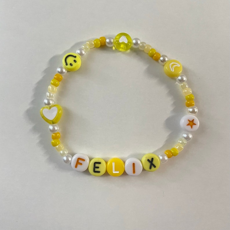 May include: A yellow and white beaded bracelet with the name "FELIX" spelled out in white letters on yellow beads. The bracelet also features a smiley face, a heart, a crescent moon, and a star.