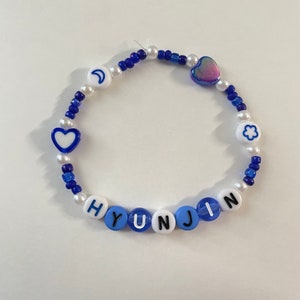 May include: A blue and white beaded bracelet with the name "HYUNJIN" spelled out in black letters. The bracelet also features a crescent moon, a heart, a flower, and a iridescent heart charm.