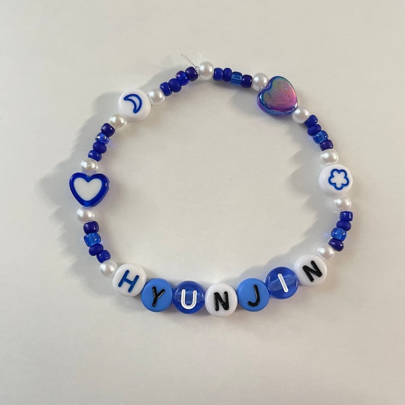 May include: A blue and white beaded bracelet with the name "HYUNJIN" spelled out in black letters. The bracelet also features a crescent moon, a heart, a flower, and a iridescent heart charm.