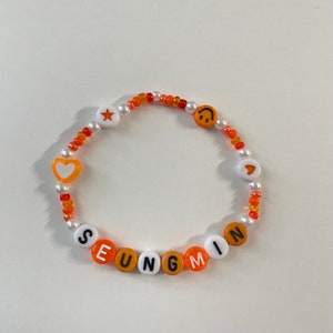 May include: Orange and white beaded bracelet with the name "SEUNGMIN" spelled out in white beads. The bracelet also features a star, heart, and smiley face charm.
